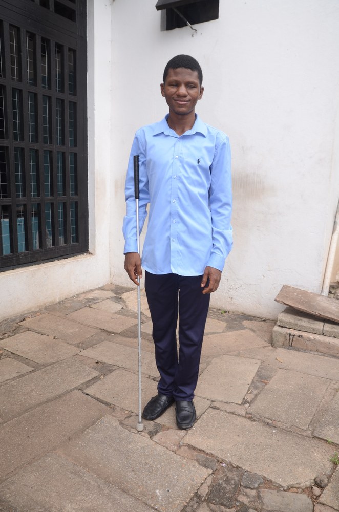 Archebold Degraft Acquah, founder of Inclusive App Craft, standing confidently in a light blue button-down shirt and dark trousers, holding his white cane against a neutral light background.